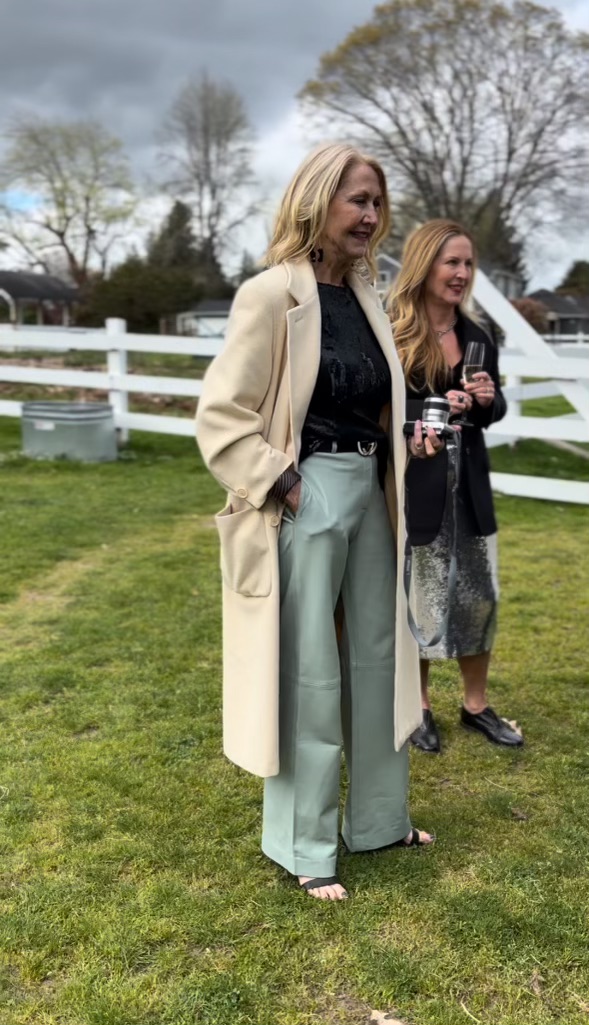 Two women in a camel coat and sage trousers on a bright lawn